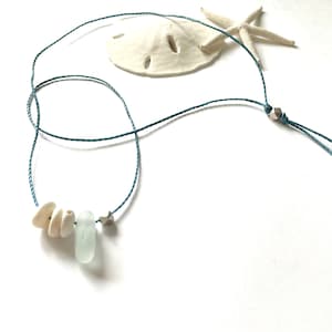 May include: A blue cord necklace with a silver bead and a charm featuring a small star. The necklace also has a sea glass bead and three white seashells.