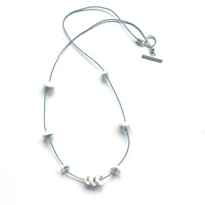 May include: A light blue cord necklace featuring several small, white, circular shell-like beads spaced along the length, with a cluster of three beads at the bottom and a silver-toned metal toggle clasp closure at the top.