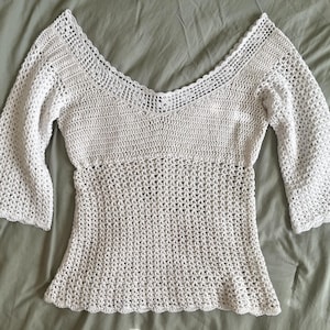 May include: A white crochet top featuring a deep V-neckline with a decorative scalloped border, three-quarter length bell sleeves, and a fitted bodice that transitions into an open, airy mesh stitch pattern throughout the lower section of the garment.