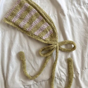 Puede incluir: Un gorro tejido en verde oliva con rayas horizontales de color rosa claro. El gorro tiene lazos largos atados con un lazo. Los extremos de los lazos son redondeados. El gorro está sobre una superficie blanca.