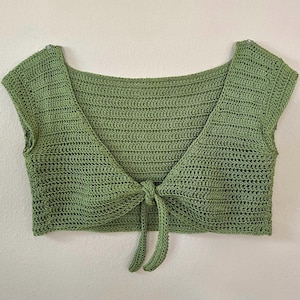 Op de afbeelding: Een groene gehaakte crop top met een vierkante halslijn en korte mouwen. De top wordt aan de voorkant vastgemaakt, waardoor een flatterend silhouet ontstaat. De handgemaakte top is een stijlvolle toevoeging aan elke garderobe.