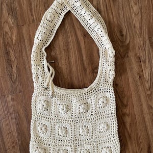 Könnte beinhalten: Cremefarbene Häkeltasche mit Blumenmuster. Die Tasche hat einen langen Schulterriemen und einen Kordelzugverschluss. Das Häkelmuster besteht aus Quadraten mit einem Blumenmotiv. Die Tasche ist aus weichem, strukturiertem Garn gefertigt.