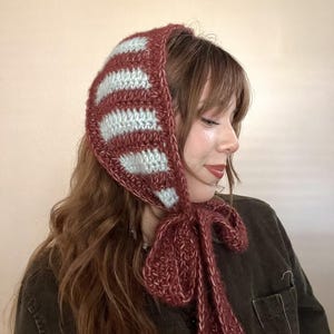 Puede incluir: Un gorro de ganchillo a rayas burdeos y azul claro con lazos largos. El gorro está hecho de un hilo suave y texturizado y está diseñado para usarse en la cabeza. Los lazos se pueden atar con un lazo debajo de la barbilla. Un accesorio hecho a mano y acogedor.