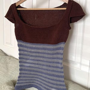 Könnte beinhalten: Ein gestricktes Oberteil mit rundem Ausschnitt und kurzen Ärmeln. Das Oberteil hat ein dunkelbraunes Oberteil und einen gestreiften blau-grauen unteren Teil. Das Kleidungsstück wird auf einem Holzbügel vor einer weißen Tür präsentiert.