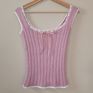 May include: A pink ribbed knit tank top with a square neckline and a delicate bow. The top features white lace trim along the neckline and armholes. The tank top is sleeveless and hangs on a wooden hanger.