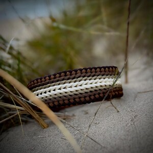 May include: A woven bracelet in brown, beige, and white, lying on sand. The bracelet has a braided design with a colour gradient, going from dark brown to light beige and white. The background shows blurred green and brown grass.