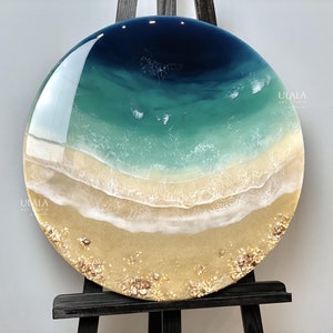 May include: A round resin art piece depicting a beach scene with blue and green water, white waves, and a sandy beach with gold glitter and seashells. The art piece is on a black easel and has the text "ULALA ART STUDIO" in the upper right corner.