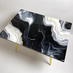 May include: A black and white coffee table with gold legs. The table top has a marbled design with black, white, and gold swirls.