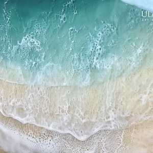 May include: A close-up of a resin art piece depicting a turquoise ocean wave crashing onto a sandy beach. The wave has a white foamy crest and the sand is a light beige color.