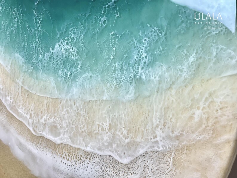 May include: A close-up of a resin art piece depicting a turquoise ocean wave crashing onto a sandy beach. The wave has a white foamy crest and the sand is a light beige color.