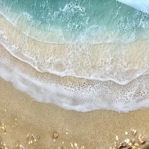 May include: Close-up of a resin art piece depicting a beach scene with a turquoise blue ocean, white foamy waves, and a sandy beach with gold glitter.