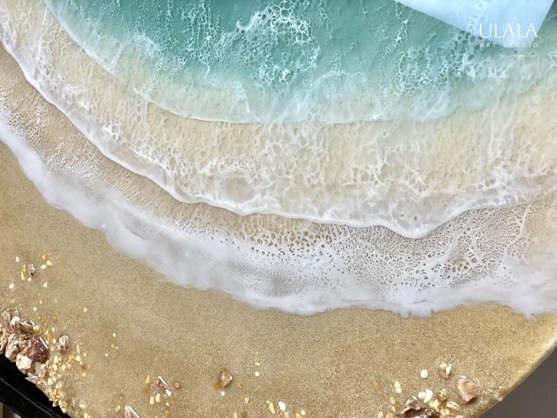 May include: Close-up of a resin art piece depicting a beach scene with a turquoise blue ocean, white foamy waves, and a sandy beach with gold glitter.