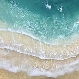 May include: A close-up of a resin art piece depicting a turquoise ocean with white foamy waves crashing on a sandy beach. The piece is circular and has a textured surface.