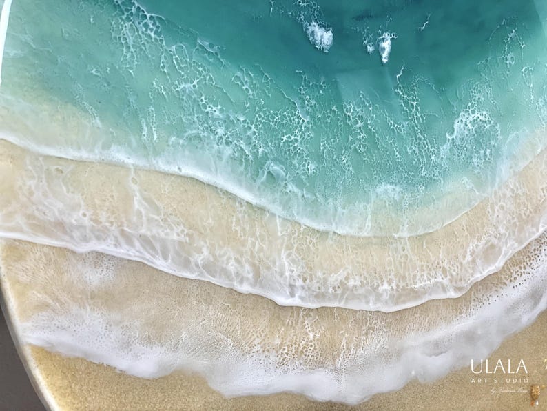 May include: A close-up of a resin art piece depicting a turquoise ocean with white foamy waves crashing on a sandy beach. The piece is circular and has a textured surface.