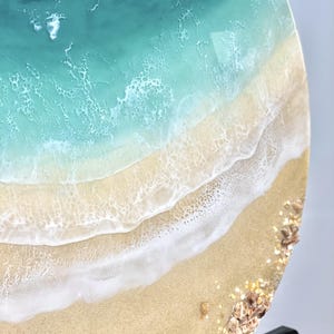May include: A close-up of a round resin art piece depicting a beach scene. The top half of the piece is a turquoise blue ocean with white foam. The bottom half is a sandy beach with white foam waves and small brown and gold shells.