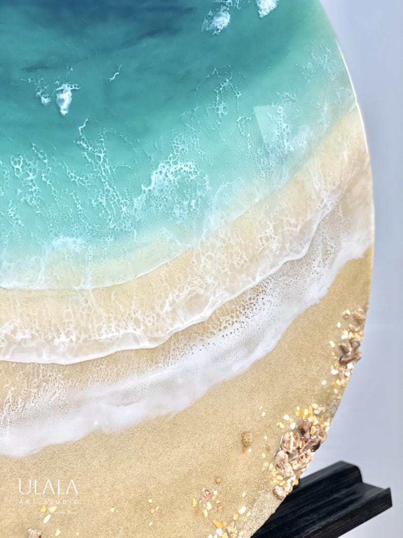 May include: A close-up of a round resin art piece depicting a beach scene. The top half of the piece is a turquoise blue ocean with white foam. The bottom half is a sandy beach with white foam waves and small brown and gold shells.