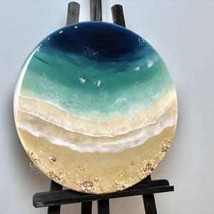 May include: A round resin art piece depicting an aerial view of an ocean beach scene. The ocean is a deep blue color, transitioning to a turquoise green color. The beach is a light tan color with white foamy waves and small white and gold accents.