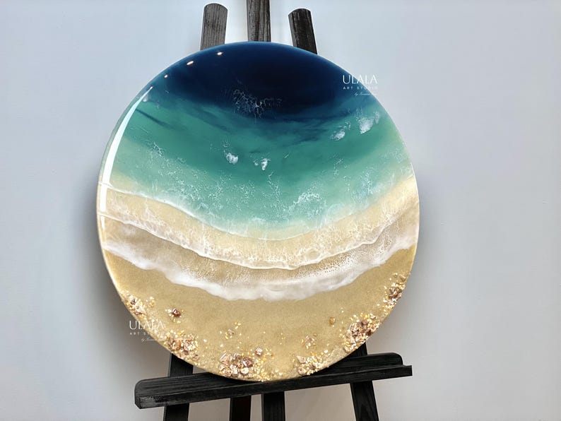 May include: A round resin art piece depicting an aerial view of an ocean beach scene. The ocean is a deep blue color, transitioning to a turquoise green color. The beach is a light tan color with white foamy waves and small white and gold accents.