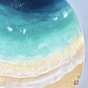 May include: A circular resin art piece depicting a beach scene with blue ocean waves crashing on a sandy shore. The ocean is a deep blue at the top, fading to a lighter blue and then turquoise. The sand is a light beige color with white foam from the waves.