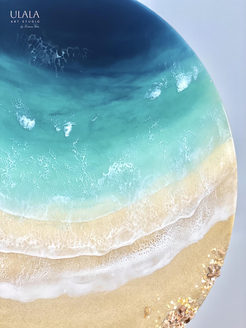 May include: A circular resin art piece depicting a beach scene with blue ocean waves crashing on a sandy shore. The ocean is a deep blue at the top, fading to a lighter blue and then turquoise. The sand is a light beige color with white foam from the waves.