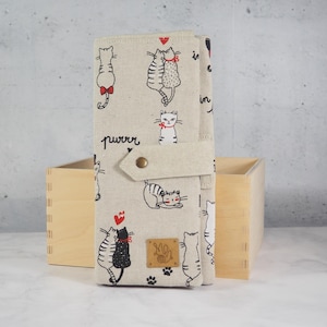 Könnte beinhalten: Eine beigefarbene Stoffbrieftasche mit Katzenmuster, die verschiedene Katzenillustrationen mit roten Schleifen und Herzen zeigt. Das Wort "purrr" ist auf der Brieftasche aufgedruckt. Die Brieftasche ist mit einem beigen Riemen verschlossen.