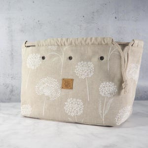Könnte beinhalten: Eine beigefarbene Stofftasche mit weißem Pusteblumenmuster, einem Kordelzug mit dicken Seilen und drei Metallösen an der Oberkante. Ein kleines, quadratisches braunes Lederetikett mit den Buchstaben MG ist mittig auf der Vorderseite angebracht.