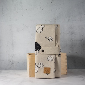 Op de afbeelding: Een beige stoffen aankleedkussen met een zwart-wit schapenpatroon. De mat is opgevouwen en vastgezet met een riem en knoop. Een kleine houten kist staat op de achtergrond. Een klein bruin leren label is bevestigd.