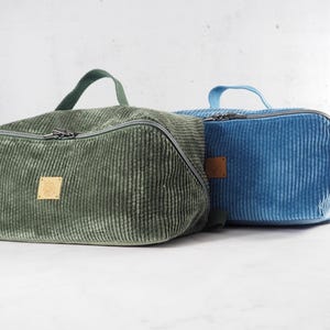 May include: Two rectangular corduroy toiletry bags, one in sage green and one in bright blue. Each bag features a top handle, a silver-toned zipper closure, and a small square leather-like patch centered on the front side of the textured fabric.