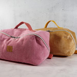 Può includere: Due borse da viaggio in velluto a coste. Una è color rosa antico, l'altra giallo senape. Entrambe hanno una forma rettangolare con la parte superiore curva, chiusura con cerniera e maniglia superiore. Le borse sono su una superficie di marmo bianco.