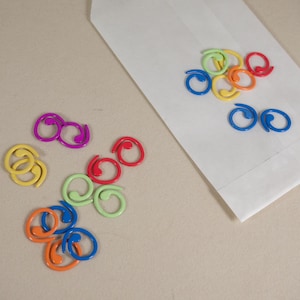 Könnte beinhalten: Eine Sammlung bunter Metall-Binderclips in verschiedenen Formen und Größen. Die Clips sind in einem Muster auf einer weißen Oberfläche angeordnet.