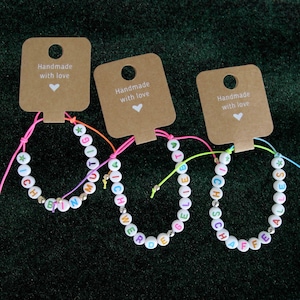 Könnte beinhalten: Drei bunte Freundschaftsarmbänder mit weißen Perlen und bunten Buchstaben, die "ICH BIN", "WERDE GELIEBT" und "ALLES SCHÖFF" buchstabieren, mit einem braunen Etikett, auf dem "Handmade with love" steht, das an jedes Armband befestigt ist.