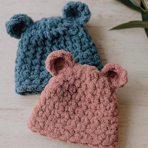May include: Two handmade crochet hats with bear ears. One hat is a dusty rose color, and the other is a teal blue. The hats are made of a soft, textured yarn. The hats are likely for babies or young children.