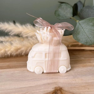Peut inclure: Une figurine en céramique blanche représentant un van vintage avec un ruban rose et un bouquet de fleurs blanches sur le dessus. Le van est posé sur une surface en bois.