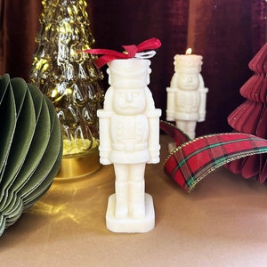 Puede incluir: Vela de cascanueces blanca con un lazo rojo, de pie sobre una superficie marrón con un árbol de Navidad dorado y una cinta de cuadros rojos y verdes en el fondo.