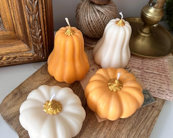 May include: Four pumpkin-shaped candles in orange and off-white colors. Two candles are tall and gourd-shaped, while two are round and squat. Each candle features a metallic gold-painted stem and a single white wick centered at the top.