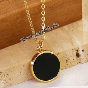 Może przedstawiać: Złoty łańcuszek z okrągłym czarnym wisiorkiem. Wisiorek ma złotą obwódkę.