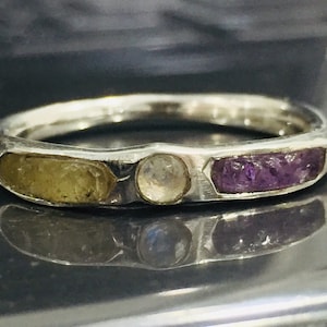 Könnte beinhalten: Ein silberner Ring mit drei darin gesetzten Steinen. Die Steine sind hellgelb, klarweiß und violetter Amethyst.