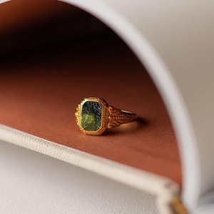 Op de afbeelding: Een gouden ring met een groene edelsteen in een vierkante zetting. De ring heeft ingewikkelde details op de band.
