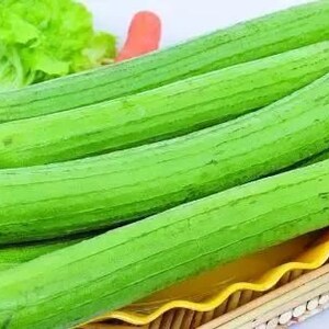 May include: A close-up of a basket filled with several long, green, ribbed loofah gourds. The gourds are arranged in a row, with the ends of the gourds slightly overlapping.