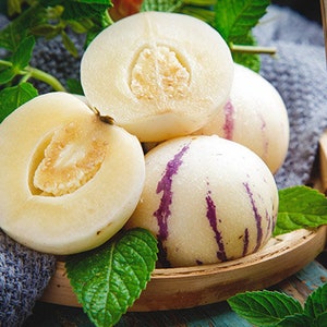 Puede incluir: Un grupo de melones pepino maduros, algunos cortados para revelar su pulpa blanca cremosa, est&aacute;n dispuestos en una cesta tejida con hojas verdes.