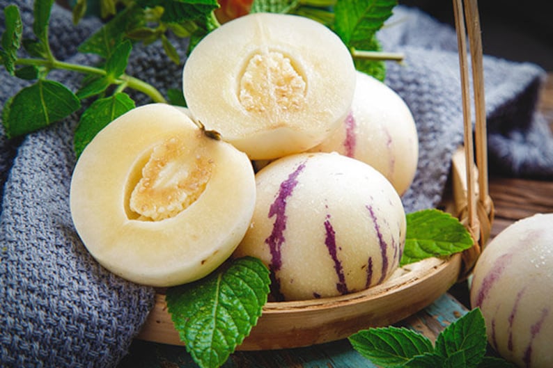 Puede incluir: Un grupo de melones pepino maduros, algunos cortados para revelar su pulpa blanca cremosa, est&aacute;n dispuestos en una cesta tejida con hojas verdes.
