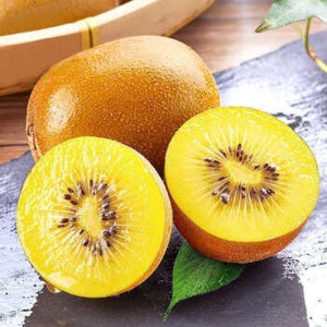 May include: Two golden kiwi fruits, one cut in half, revealing the yellow flesh and black seeds. The kiwi is on a dark gray surface with a green leaf.
