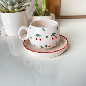 May include: White ceramic teacup and saucer set with a cherry design. The cup has a rounded shape and a handle. The saucer has a red rim.