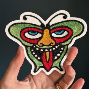 May include: A green, red, yellow, and black embroidered butterfly patch with a cartoon face. The butterfly has large eyes, a wide mouth, and a long tongue.