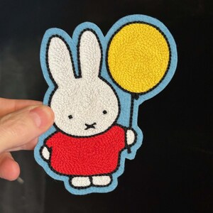 Op de afbeelding: Geborduurde patch met een witte konijn met een rood shirt en een gele ballon. Het konijn heeft zwarte ogen en een zwarte 'x' mond. De patch is blauw omlijnd.