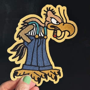 May include: An embroidered patch featuring a cartoon vulture character with a tan head, a large yellow beak, and a cigar. The character wears blue denim overalls with teal straps over a white and black striped shirt, standing on yellow feet.