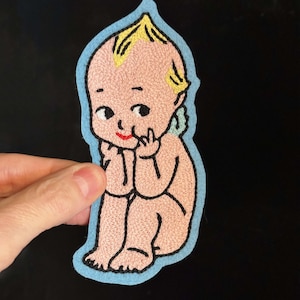 May include: Embroidered patch featuring a seated baby with pink skin, black eyes, and a red mouth. The baby has yellow hair and is outlined in light blue. The patch is likely for clothing or accessories.