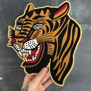 May include: Embroidered tiger head patch with a fierce expression. The patch features a tan background with black stripes, white teeth, red accents, and a yellow border. This patch is suitable for jackets, bags, or other fabric items.