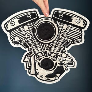 Pode incluir: Um tapete preto e branco em forma de motor de motocicleta. O tapete apresenta um design detalhado dos componentes do motor, incluindo cilindros, pistões e outras peças mecânicas. O tapete é segurado por uma mão.