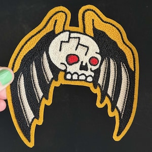 May include: An embroidered patch featuring a central white skull with a cracked forehead and bright red eyes. The skull is flanked by two large, stylized wings with black and beige striped feathers, all outlined with a thick mustard yellow border.